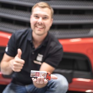 Ein Mann zeigt einen Miniatur-LKW vor einem grossen roten Fahrzeug und hebt den Daumen.