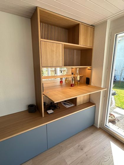Moderner Schrank aus Eichenholz mit integriertem Regal und Arbeitsfläche, darauf Flaschen und Accessoires, in einem hellen Raum mit Fenster.