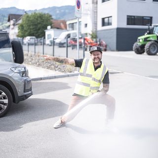 Mann in Warnweste zeigt lachend auf ein Auto.