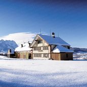 Schneebedecktes Haus in alpiner Landschaft mit klarblauem Himmel im Hintergrund.