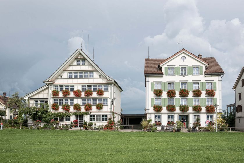Zwei traditionelle Gebäude mit blühenden Balkonen vor einem bewölkten Himmel.