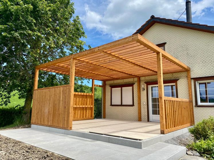 Holzterrasse mit Überdachung vor einem Haus unter blauem Himmel.