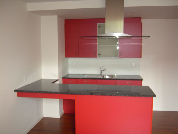 Moderne Küche mit roten Schränken, grauer Arbeitsplatte und dunklem Holzboden.
