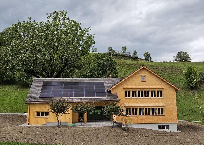 Modernes Holzhaus mit Solarpanelen vor einer grünen Hügelkulisse.