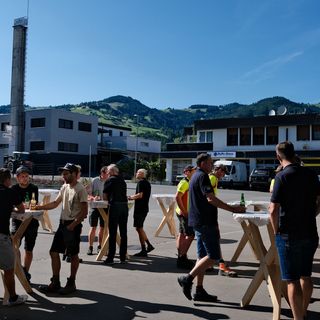 Gruppe Menschen bei einer Veranstaltung im Freien vor industrieller Kulisse mit Bergen.