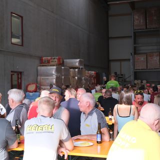 Mitarbeiter und Gäste sitzen in einer Lagerhalle von Stark AG Altstätten und geniessen ein Zusammensein an Bierbänken.