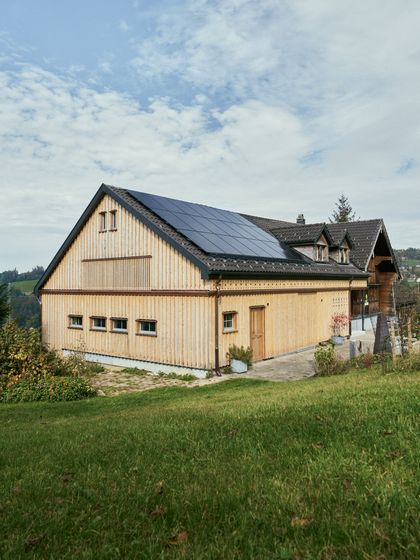 Modernes Holzhaus mit Solarpanelen auf grüner Wiese unter blauem Himmel.