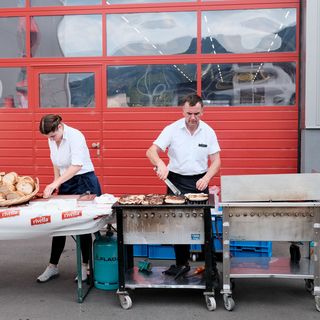 Ein Mann und eine Frau bereiten an einem Grillstand mit Rivella-Tischdecke Speisen vor.