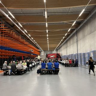Menschen sitzen an langen Tischen in einer grossen Halle der Stark AG, umgeben von Holzmaterialien.