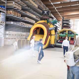 Menschen erkunden eine Lagerhalle mit aufblasbaren Spielstrukturen an einem Holzhändler-Standort.