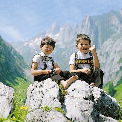 Zwei Jungen in traditioneller Tracht sitzen auf einem Felsen vor einer alpinen Kulisse.