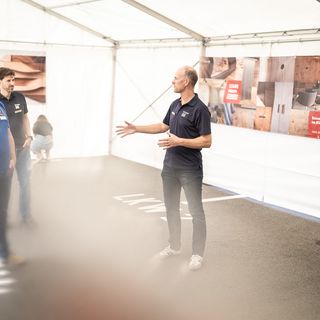 Ein Mann hält eine Präsentation vor einer kleinen Gruppe in einem Ausstellungszelt mit Holzmustern im Hintergrund.
