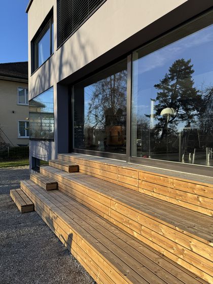 Moderne Holzterrasse mit grossen Glasfenstern in der Sonne.