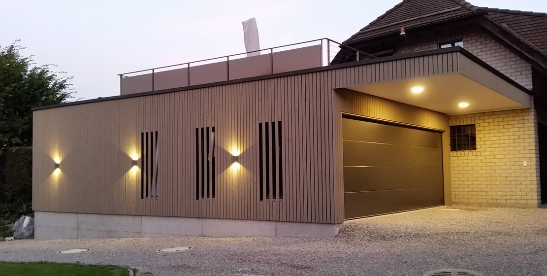 Modernes Gebäude mit beleuchteter Holzfassade und Terrasse bei Abenddämmerung.