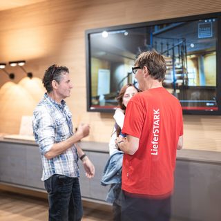 Drei Personen diskutieren in einem modernen Ausstellungsraum mit Holzverkleidung und grossem Bildschirm.