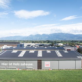 Luftaufnahme der Stark AG mit dem Slogan 'Hier ist Holz zuhause' auf der Lagerhalle vor alpiner Kulisse.