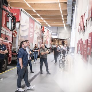 Personen betrachten Informationstafeln in einer Lagerhalle neben Lastwagen.