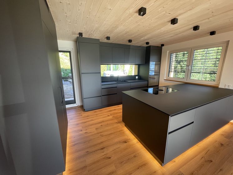 Moderne Küche mit Holzdecke und mattschwarzen Schränken, beleuchteten Böden und grossem Fenster.