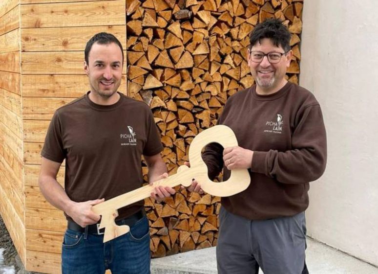 Zwei Männer halten einen grossen hölzernen Schlüssel vor einem Stapel Holz.
