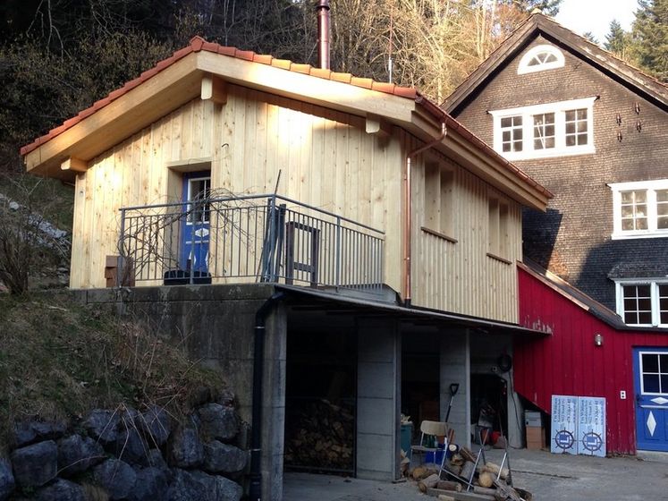 Modernes Holzgebäude auf einem Hang mit darunterliegendem Garagenbereich, umgeben von ländlicher Architektur.