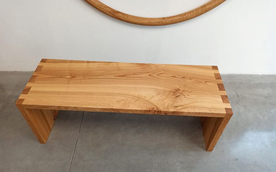 Minimalistische Holzbank aus hellem Holz mit Zinkungen auf Betonboden.