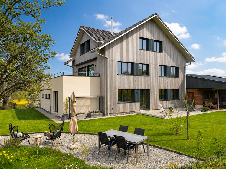 Modernes Einfamilienhaus mit Holzfassade und gepflegtem Garten bei strahlendem Sonnenschein.