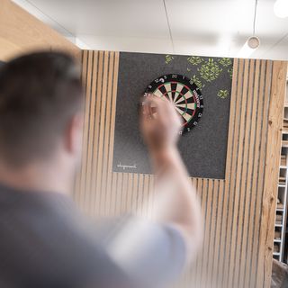Person wirft Dartpfeil auf Dartscheibe in modernem Büroumfeld.