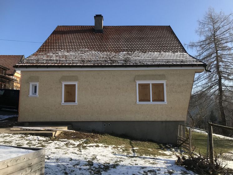 Haus mit schneebedecktem Dach in ländlicher Umgebung, viele Fenster mit Holzläden.