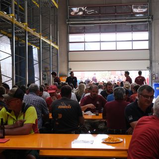 Menschen sitzen bei einem Firmenevent in einer Lagerhalle an Biertischen.