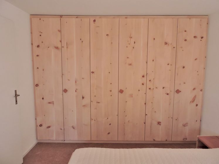 Ein mehrtüriger Schrank aus naturbelassenem Holz mit sichtbaren Astknoten in einem minimalistisch eingerichteten Raum.