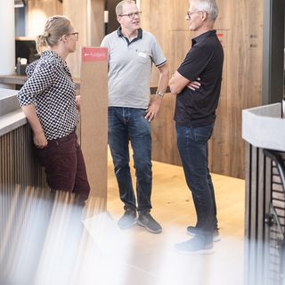 Drei Personen unterhalten sich in einem modern gestalteten Ausstellungsraum mit Holzverkleidung.